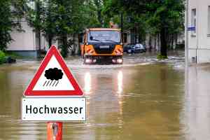 © Animaflora PicsStock - Adobe Stock.com Hochwasserschild vor überfluteter Straße, Straßenmeisterfahrzeug im Hintergrund